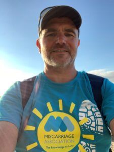 Man in cap with Miscarriage UK Miles That Matter t-shirt on, with blue sky behind him.